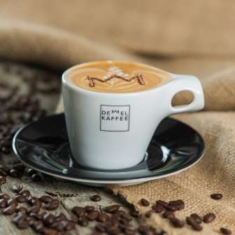 Cappuccino with crown in the foam in a cup of Demmel coffee, surrounded by coffee beans on wood and jute sack - enjoyment in Liechtenstein
