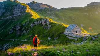 Hiker looks at the Pfälzer hut in Liechtenstein, surrounded by a green mountain landscape and Alpine panorama in the evening light