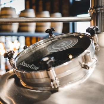 Close-up of a round viewing window on a shiny brewing kettle with a view of the brewing hall in the background.
