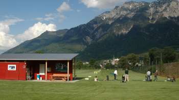 Golfers on the driving range in Liechtenstein with a view of green meadows, a red clubhouse and the surrounding mountain landscape under a blue sky.