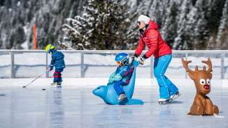 Mother pushes child on an ice skating penguin across the ice rink in Malbun, in the background a child with an ice hockey stick