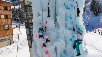 Two children ice climbing on the frozen climbing tower in Malbun with ice axes and helmets