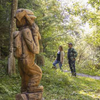 Two hikers discover a carved figure on the Walser Saga Trail in the middle of a light-flooded forest