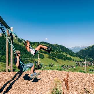 Two children swinging on a mountain playground with a view over the summer panorama of Malbun in Liechtenstein