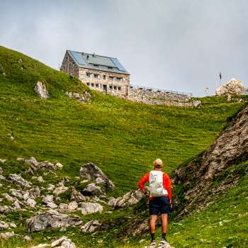 Hiker with a red rucksack looks at the Pfälzer hut in the middle of a green mountain landscape.