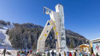 Malbun ice tower under a blue sky, surrounded by snow-covered trees and skiers