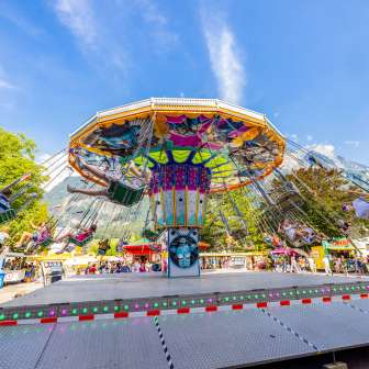 Large colorful chain carousel at the fair in Balzers, full of happy visitors in front of a mountain backdrop under a blue sky.