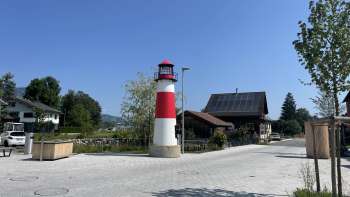 Small red and white Moby lighthouse on a square in Ruggell in sunny weather, with traditional buildings in the background.
