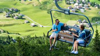 Two people on the Sareis pleasure chairlift with a view of the green valley in the background