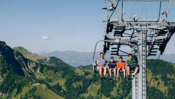 A family rides the Sareis chairlift over a green mountain landscape
