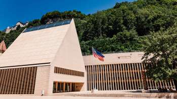 Modern parliament building in Vaduz with a pointed gable, the Liechtenstein flag in front of it and surrounded by green hills.