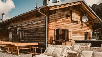 Cosy sun terrace with wooden benches and comfortable lounge furniture in front of the Sareis mountain hut at an altitude of 2,000 meters, with bright sunshine and a mountain backdrop.