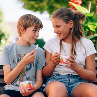 Two children sit on a park bench in Liechtenstein and eat ice cream - smiling faces, flowers and a summer atmosphere in the countryside