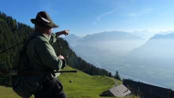 A man in traditional dress points from a mountain meadow into the Rhine Valley.