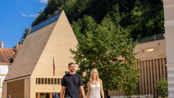 A couple strolls across the sunny Government Square in Vaduz with a view of the modern parliament building and Vaduz Castle in the background.