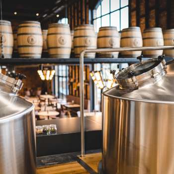 View of a modern brewery with gleaming stainless steel tanks and stacked wooden barrels in the background.