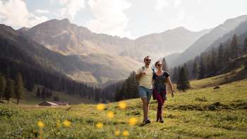 Two people hiking through a wide alpine meadow with a view of the surrounding mountains of Alp Valüna in sunny weather Two people hiking through a wide alpine meadow with a view of the surrounding mountains of Alp Valüna in sunny weather