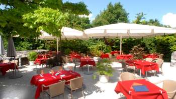 Exterior view of the terrace with red tablecloths, surrounded by trees and parasols.