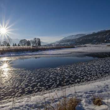 Sunrise at Lake Egelsee in Mauren in winter with glistening ice, snow and a sunny mountain backdrop.