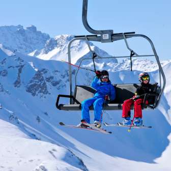 Two children with ski equipment ride in a chairlift through the snowy mountains of Malbun
