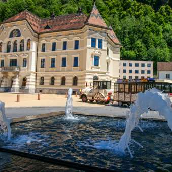 View of the government building in Vaduz with the Citytrain passing by and water fountains in the foreground