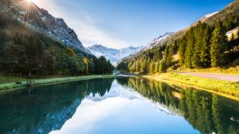 Mirror-smooth Gänglesee lake in Steg, framed by dense forests and snow-capped mountains in the sunshine