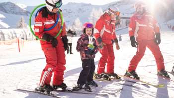 Three ski instructors in red suits and a child on skis in the snow, snow-covered mountains and sunshine in the background