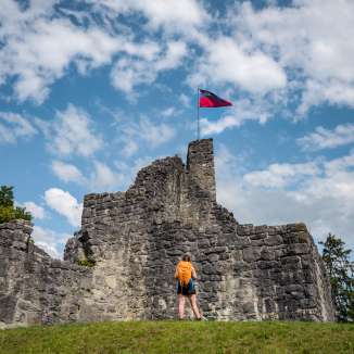 Hiker in front of the historic ruins of Schellenberg Castle with a waving Liechtenstein flag under a blue sky