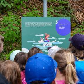 Group of children looking at an information board with an illustration of "Mila in the Wonder Tunnel" along the Gafadura Trail in the forest.