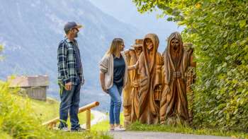 Two hikers look at carved wooden figures on the Walser Saga Trail, a themed trail in the middle of an alpine landscape with a view of the mountains