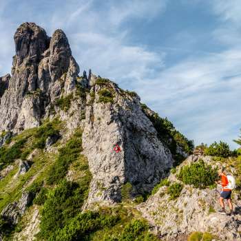 Hikers on a rocky mountain path in front of a rugged ridge with a red and white signpost