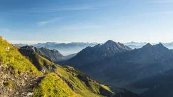 Alpine hiking trail with views of a green mountain landscape and rugged peaks in the morning sun.