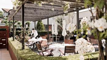 An elegant terrace with white tables set with napkins and glasses under a wooden pavilion, surrounded by lush greenery.