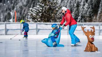 Family at the ice rink during Schlucher-Treff Malbun, supported by an ice skating aid in the form of a blue animal