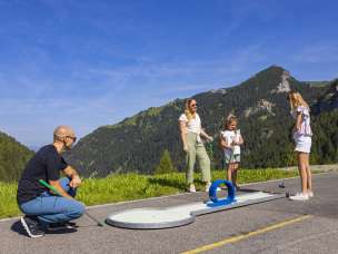 A family enjoys a round of mini-golf in sunny Malbun, Liechtenstein, surrounded by green Alpine scenery and mountains in the summer.