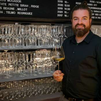 Friendly bartender of the WY Weinbar with wine glass in front of a stylish shelf full of glasses and wines.