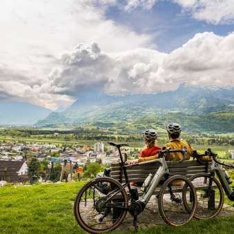 Two cyclists wearing helmets sit on a bench above the Rhine Valley and enjoy the view of the valley and the surrounding mountains - gravel bike experience in Liechtenstein