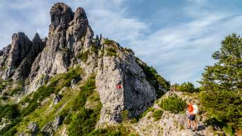 Hikers on a rocky mountain path in front of a rugged ridge with a red and white signpost
