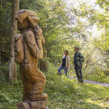 Two hikers discover a carved figure on the Walser Saga Trail in the middle of a light-flooded forest