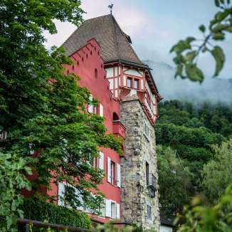Historic Red House in Vaduz, embedded in green nature with a view of the surrounding mountains and a slightly cloudy sky.