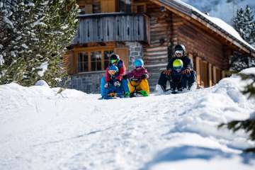 Two adults and three children sit on sledges and start a descent together in front of a snow-covered mountain hut in Malbun
