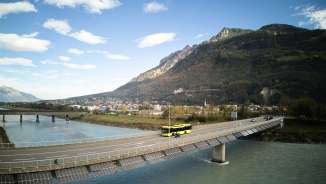 Yellow LIEmobil bus crosses the Rhine bridge near Vaduz with a mountain panorama in the background - Experience Liechtenstein