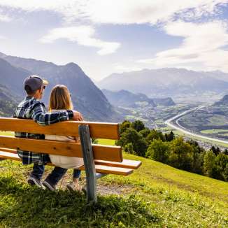 Couple sitting on a bench in Triesenberg looking out over the Rhine Valley in Liechtenstein with an impressive Alpine panorama