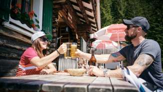 Two people clink glasses of beer in front of the Gafadura hut in the Liechtenstein Alps