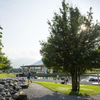 Sunny walk with shady trees and a view of the water in the Grossabünt recreational park in Ruggell.