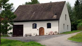 Traditional mill in Eschen with a white façade, dark shutters and an old millstone ensemble in front of the entrance - surrounded by green meadows and trees.