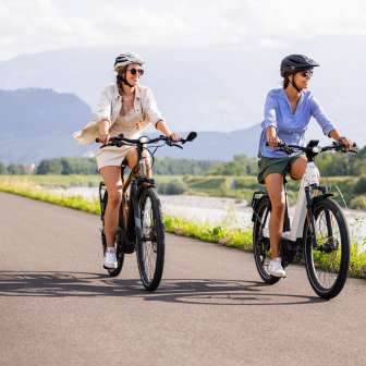 Two women on e-bikes on the Rhine cycle path near Liechtenstein, with river, green landscape and mountains in the background