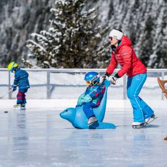 Mother pushes child on an ice skating penguin across the ice rink in Malbun, in the background a child with an ice hockey stick