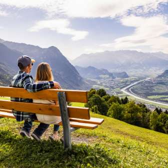 Couple sitting on a bench in Triesenberg looking out over the Rhine Valley in Liechtenstein with an impressive Alpine panorama