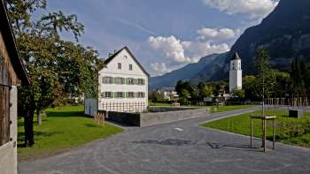 Historic vicarage with a white façade and green shutters in the middle of an open square, with the church of Balzers and the surrounding mountain landscape in the background.